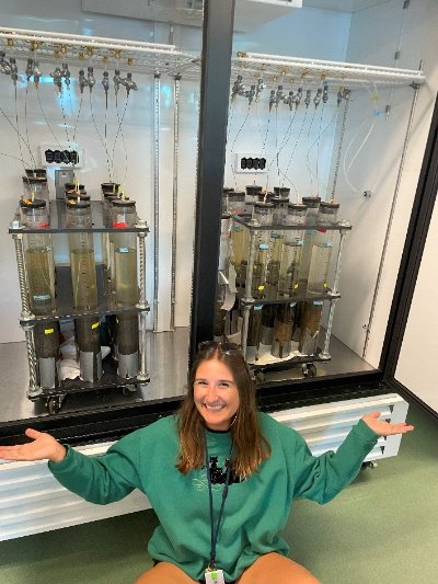 Kate Lucas poses by two racks of sediment cores in a large incubator.